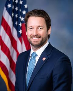 Joe Cunningham standing in front of an American flag, wearing a blue suit and light blue tie and smiling